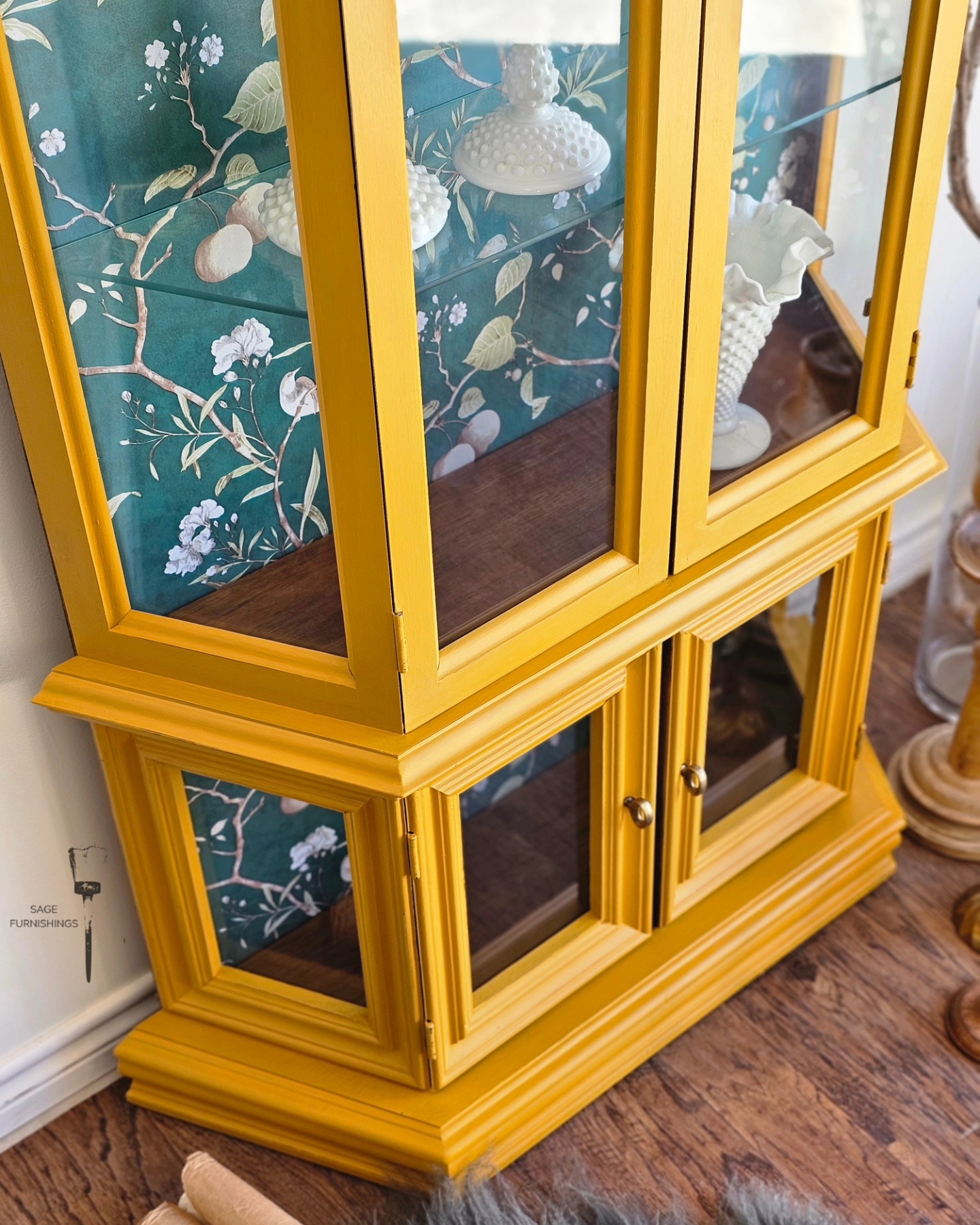 Mustard yellow painted curio cabinet with dark green floral wallpaper backing